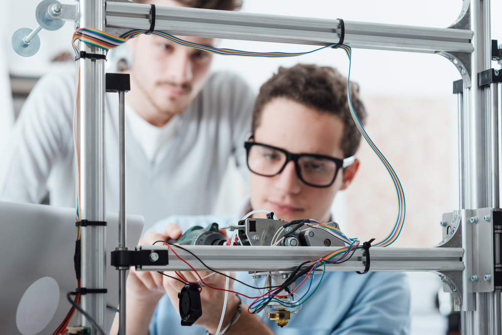 Young engineer in the lab adjusting a 3D printer's components, a student is watching